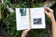 Hands holding an open gardening book: "Prune Indoor Herbs" & "Use Cloches on Greens for Cold Weather Protection." Lush green plants surround it.