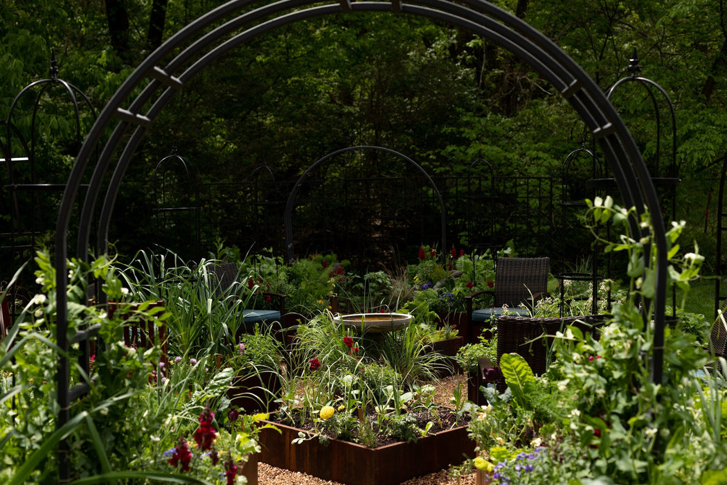 Garden archway frames raised beds with colorful plants, flowers, and a bird bath. Outdoor chairs and lush greenery in background.