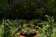 Garden archway frames raised beds with colorful plants, flowers, and a bird bath. Outdoor chairs and lush greenery in background.
