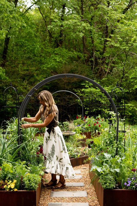 Woman tending vibrant raised garden beds with climbing plants on a trellis. Lush greens, flowers, gravel path, and forest backdrop.