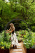 Woman tending vibrant raised garden beds with climbing plants on a trellis. Lush greens, flowers, gravel path, and forest backdrop.