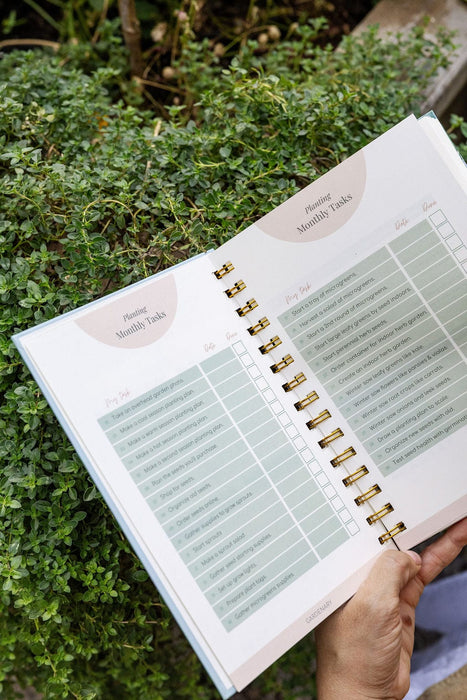 Open gardening planner displays "Planting Monthly Tasks" for seed starting, microgreens, and garden planning, held over green foliage.