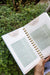 Open gardening planner displays "Planting Monthly Tasks" for seed starting, microgreens, and garden planning, held over green foliage.