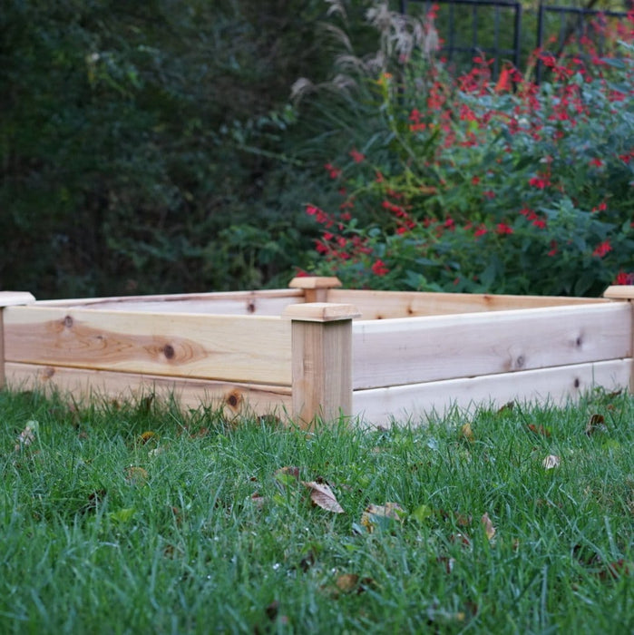 Square wooden raised garden bed on green lawn. Ready for planting. Red flowers and lush greenery in backyard.