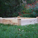 Square wooden raised garden bed on green lawn. Ready for planting. Red flowers and lush greenery in backyard.