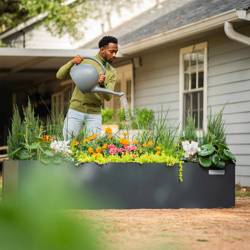 Man waters colorful plants in a dark gray Vego Garden raised garden bed. Modern outdoor planter for vibrant flowers.