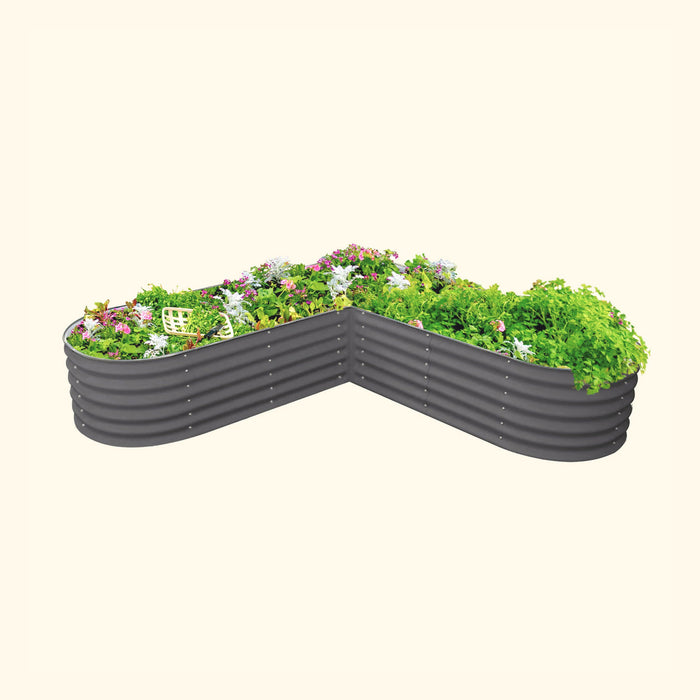 Angled dark grey corrugated steel raised garden bed with diverse green plants, pink and white flowers, and a gardening tool.