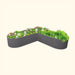 Angled dark grey corrugated steel raised garden bed with diverse green plants, pink and white flowers, and a gardening tool.