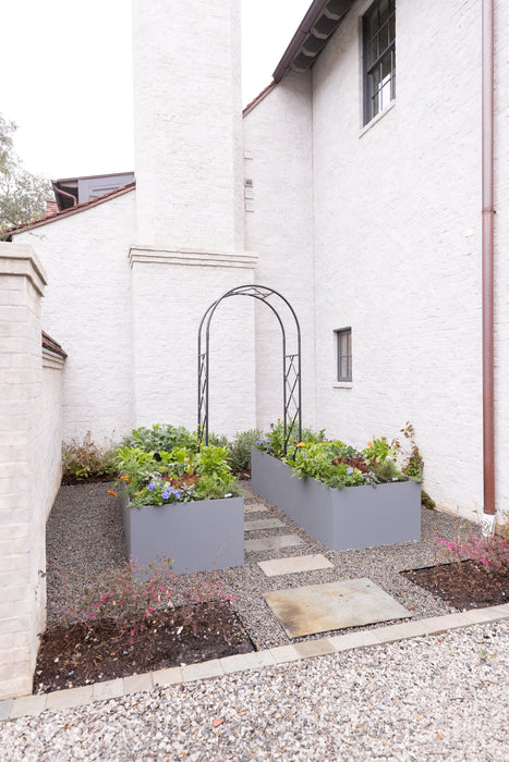 Contemporary edible garden featuring gray raised beds, lush plants, a black arch, gravel path, and white brick house.