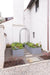 Contemporary edible garden featuring gray raised beds, lush plants, a black arch, gravel path, and white brick house.
