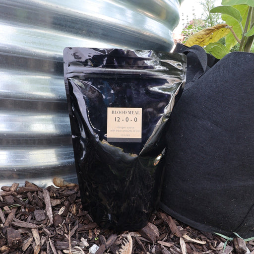 Black bag of Blood Meal 12-0-0 nitrogen fertilizer on mulch, beside a metal raised bed & fabric grow bag.