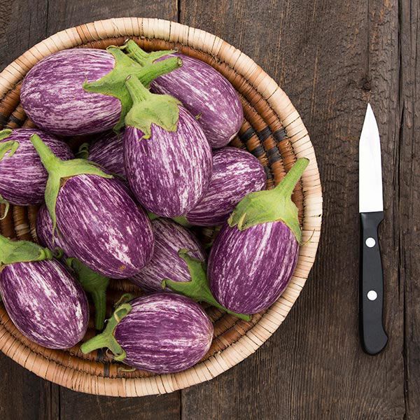 Fresh purple and white striped graffiti eggplants in a wicker basket on a rustic wooden table with a knife.