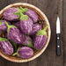 Fresh purple and white striped graffiti eggplants in a wicker basket on a rustic wooden table with a knife.