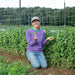 Smiling gardener holding fresh organic pea pods in a lush garden with a pea trellis.
