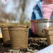 Biodegradable peat pots filled with potting soil on a wooden surface, ready for planting. Child gardening in background.
