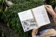 Open gardening planner showing cool season planting guide and garden layout, held over thriving green plants.
