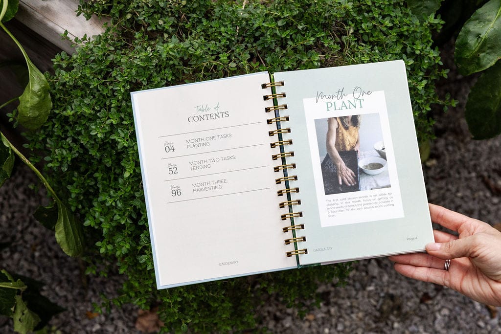 Open garden journal with planting, tending, harvesting sections. A hand holds it over lush green plants like thyme.