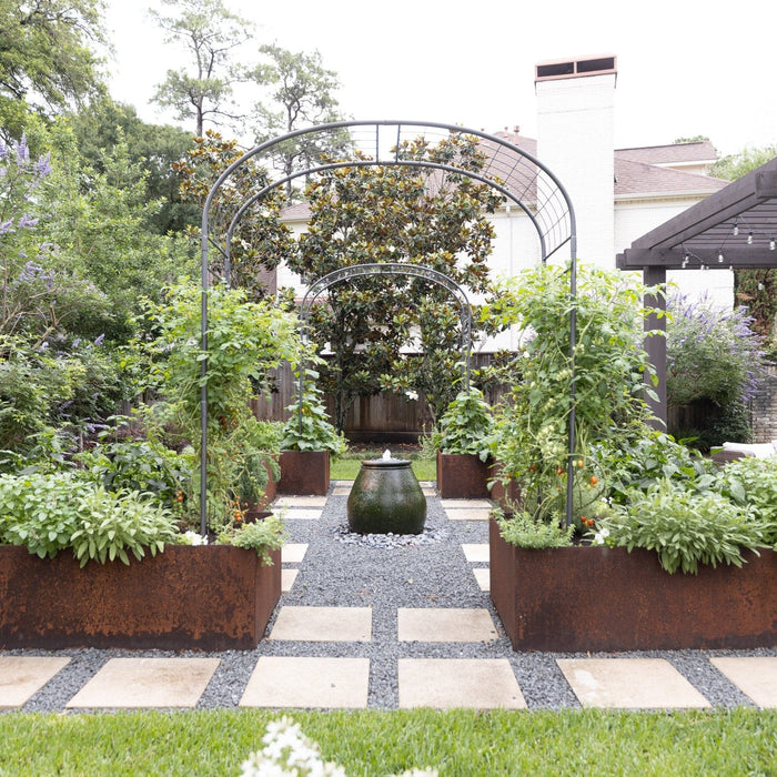 Modern backyard garden with corten steel raised vegetable beds, tomato plants, herbs, central fountain, gravel path, and pergola.