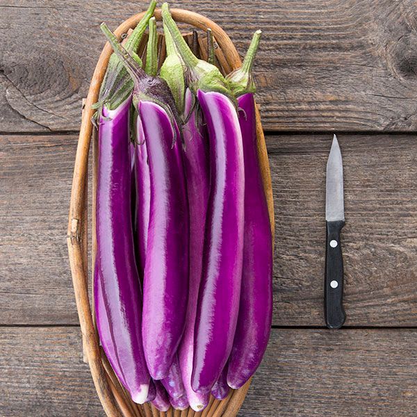 Fresh long purple Asian eggplants in a basket with a knife on a rustic wooden table.