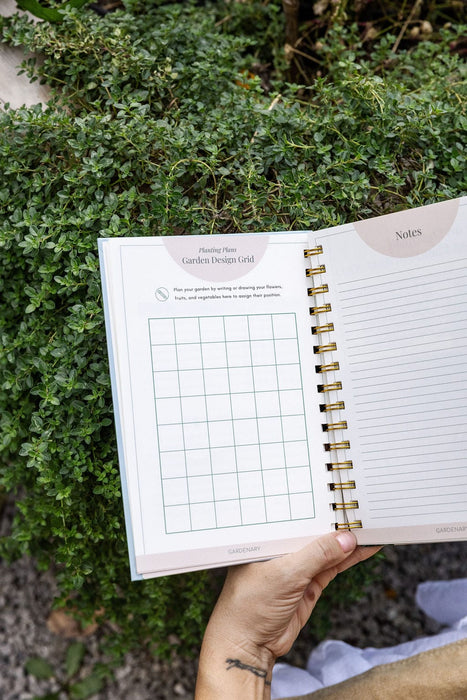 Gardenary garden design grid planner for planting plans and notes, held over lush green plants.