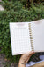 Gardenary garden design grid planner for planting plans and notes, held over lush green plants.
