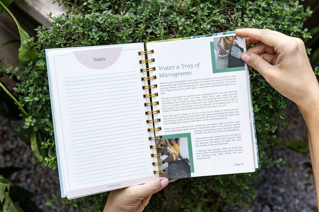 Hands hold a Gardenary microgreens growing guide open to watering instructions. Green plants in background.