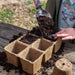 Child's hand scoops potting soil into biodegradable seed starter pots on a wooden table. Preparing for spring gardening & planting.