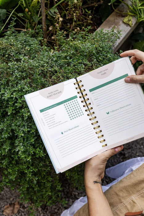 Open garden planner with weekly habit tracker, goals, pest, and pollinator observation pages held by gardener among green plants.