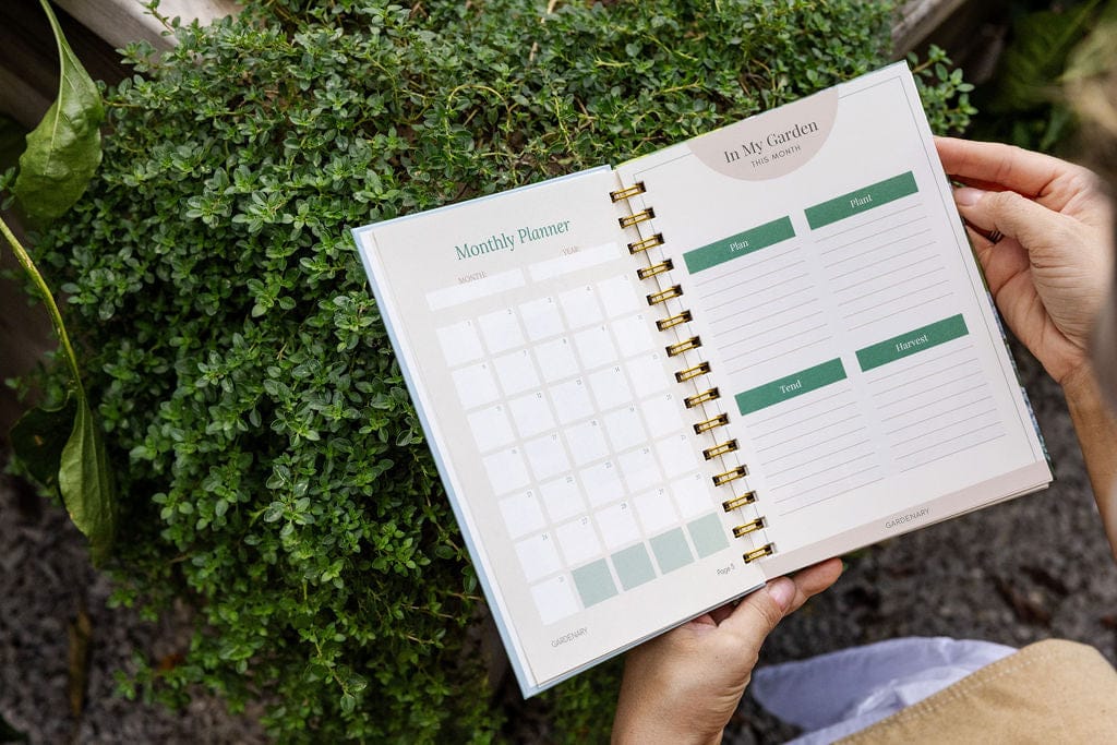 Hands hold a spiral "In My Garden" monthly planner with plant/harvest sections, over vibrant green herbs.