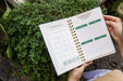 Hands hold a spiral "In My Garden" monthly planner with plant/harvest sections, over vibrant green herbs.