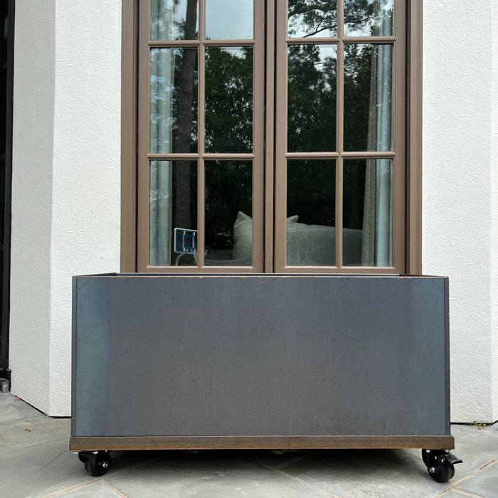 Corten steel mobile planter box on casters, placed by a brown window, white stucco wall, and stone patio.