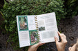 Hands hold Gardenary book open to seed ordering and sprout growing pages. Features carrots, beets, and sprout supplies. Lush garden background.