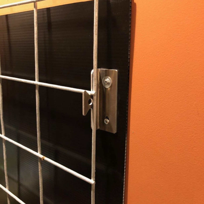 Silver metal mounting bracket securing a wire grid panel to a black board, attached to an orange wall.