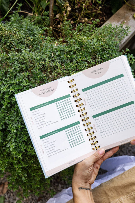 Hand holding a "Weekly Garden Habit Tracker" with indoor garden tasks like microgreens, seedlings, and goals, amidst green plants.