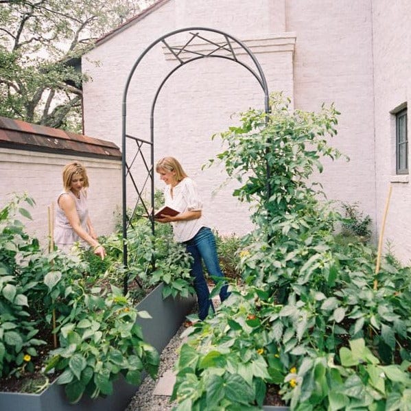 Women tending a vibrant raised vegetable garden with a metal arbor. One reads, the other examines plants.