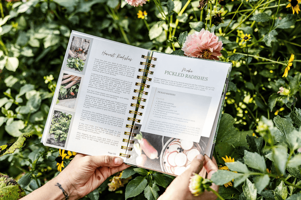 Hands hold a gardening cookbook featuring radish harvest and pickled radish recipes, surrounded by vibrant garden flowers.