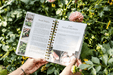 Hands hold a gardening cookbook featuring radish harvest and pickled radish recipes, surrounded by vibrant garden flowers.