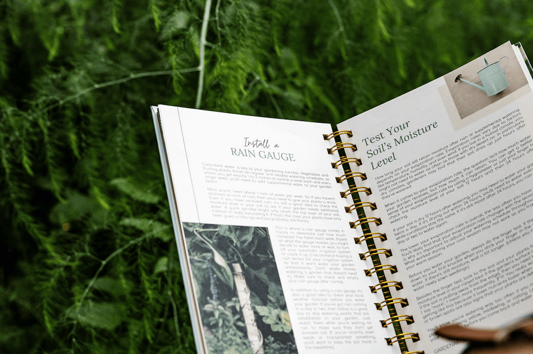 Gardening book open to watering tips: "Install a Rain Gauge" & "Test Your Soil's Moisture Level," on green foliage.