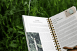 Gardening book open to watering tips: "Install a Rain Gauge" & "Test Your Soil's Moisture Level," on green foliage.