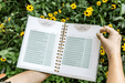 Open gardening planner showing "Planting Monthly Tasks" checklist for garden organization, held in a blooming yellow flower garden.