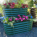 Tiered green corrugated metal raised garden bed with red coleus, green grass, pink cyclamen, blue pansies. Stone fountain background.