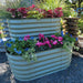 Two-tiered sage green corrugated metal garden planter with red coleus, green grass, pink cyclamen, blue pansies.