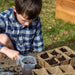 Child planting seeds in biodegradable starter trays with soil & water outdoors. Young gardener learning spring planting.