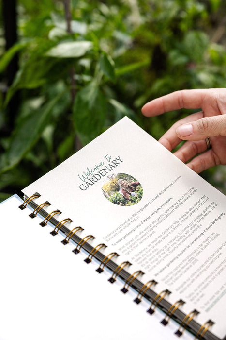 Hand holds Gardenary gardening planner, "Welcome" page featuring garden coach Nicole Johnsey. Green garden backdrop.