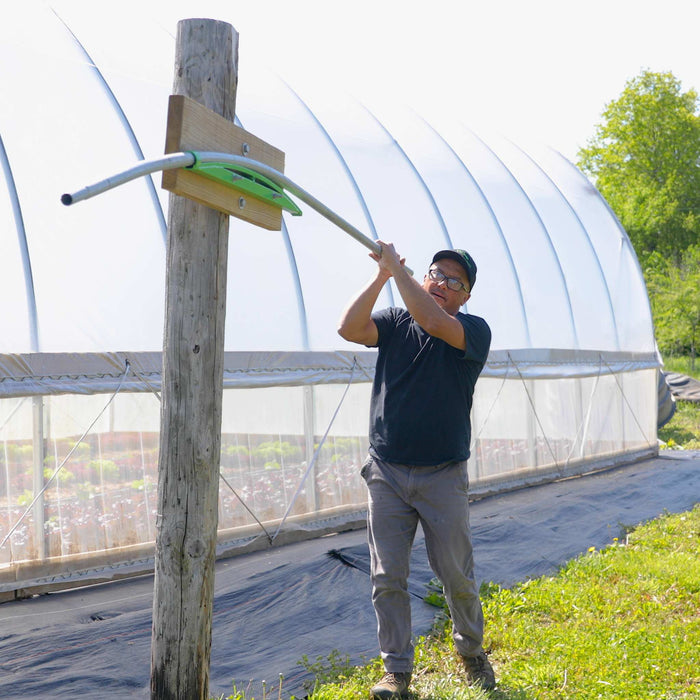 Farmer adjusts high tunnel greenhouse ventilation arm. Crops growing inside farm structure.