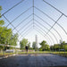Man constructing a greenhouse hoop house frame with metal arches, ladder, and black ground cover. Farm building in progress.