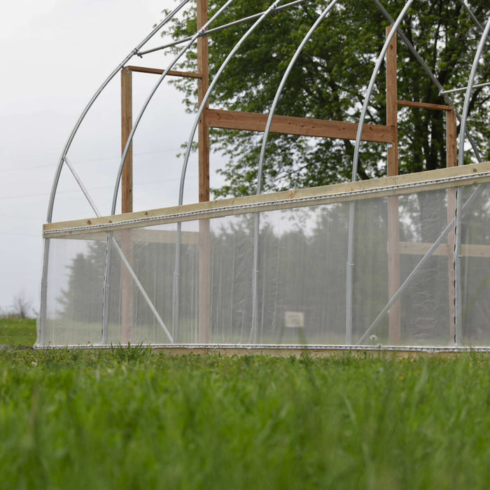 Hoop house greenhouse frame with metal arches, wood base, and clear plastic siding on a grassy field.