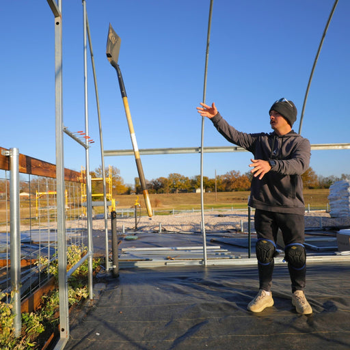Construction worker in beanie and knee pads, shovel mid-air, building a greenhouse with raised garden beds and weed barrier.