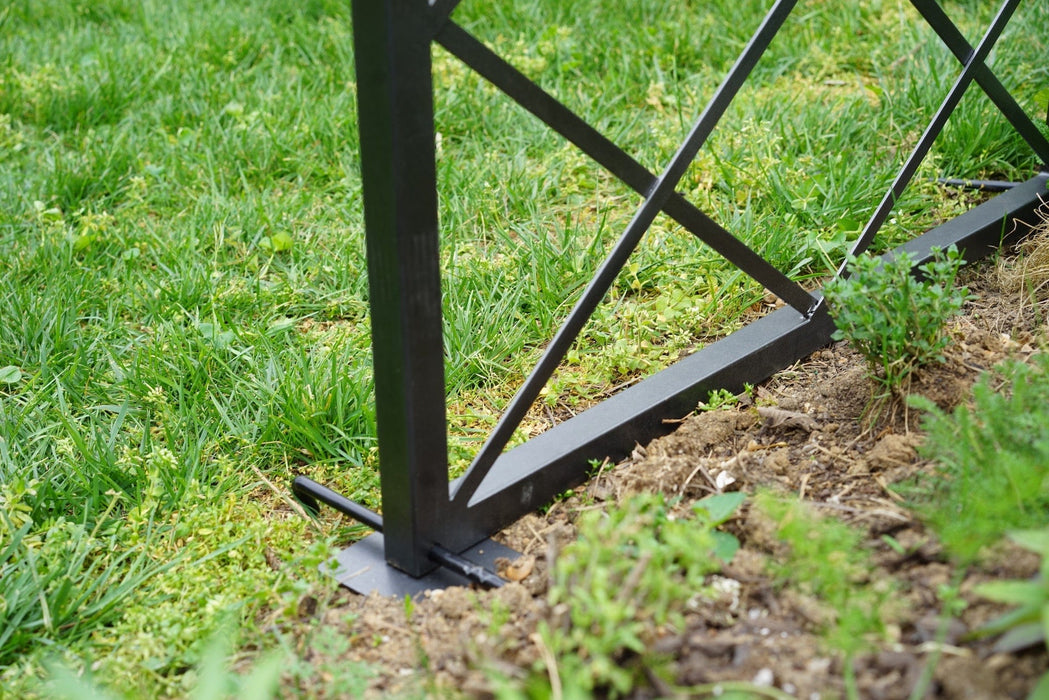 Black metal fence base with cross-braces, anchored in soil, separating green grass from a garden bed. Outdoor landscaping.