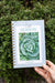 Hand holding Gardenary 'Cold Season' planner with savoy cabbage cover, against green thyme. Winter gardening journal.